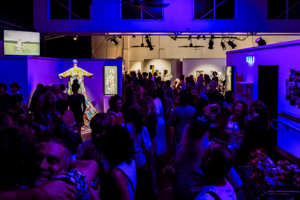 An art gallery with a crowd of people in it. You can see some sculpture of a figure like form in the distance but the people mostly obscure the view. There is a blue light on the crowd.