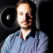 An image of a man short brown hair and some facial hair. Wearing a blue shirt and posing in front of a speaker