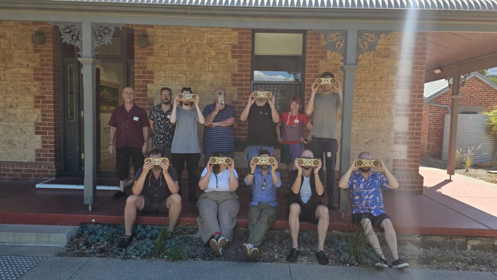 Five people seated next to each other in a row. Another five people stand behind them for the group photo. Everybody has a wooden box held up covering their face.